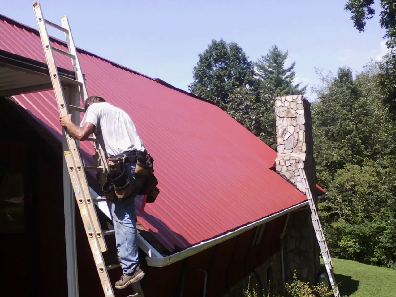 Roofer on a ladder beside a red metal roof and stone chimney — Soffit & Fascia Repair by Roofing Brothers in Camanche